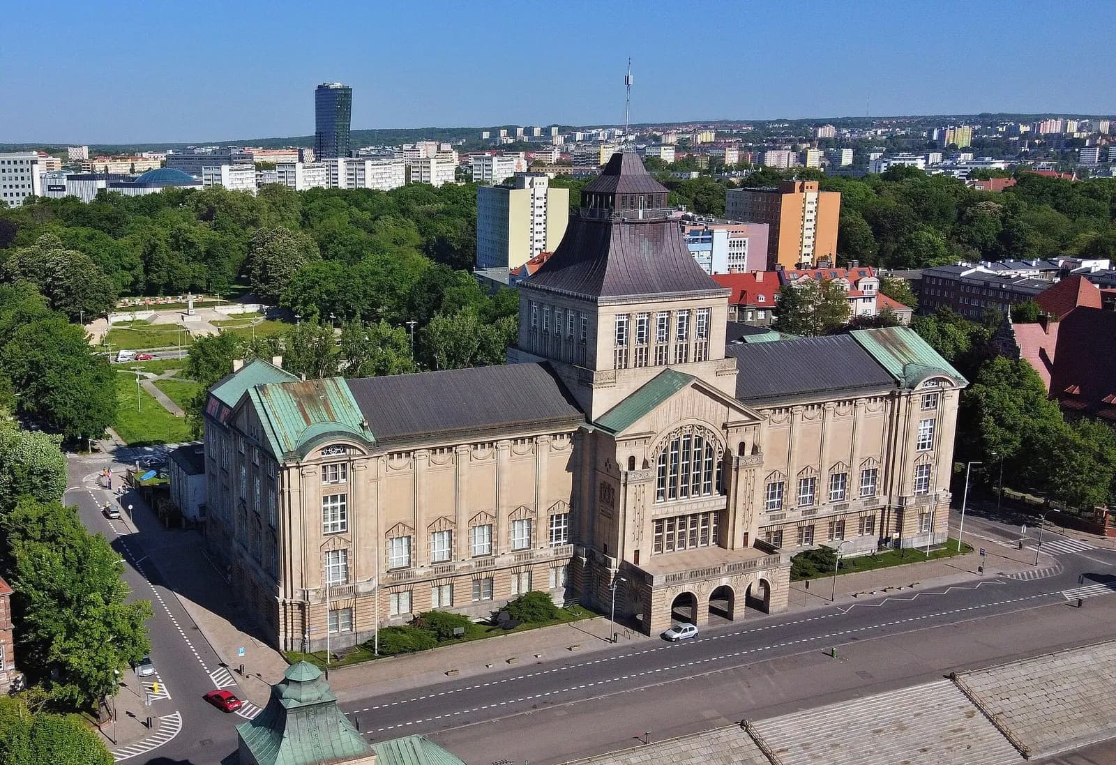 An aerial view of a large, ornate stone building with a distinctive turret and green roofs, situated on a tree-lined street in a bustling city.
